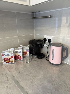 A kitchen counter with a coffee maker, two Tassimo coffee pods, a glass cup, and a kettle. - Large Private Suite for Families & Workers Near Hospital (Greater London)