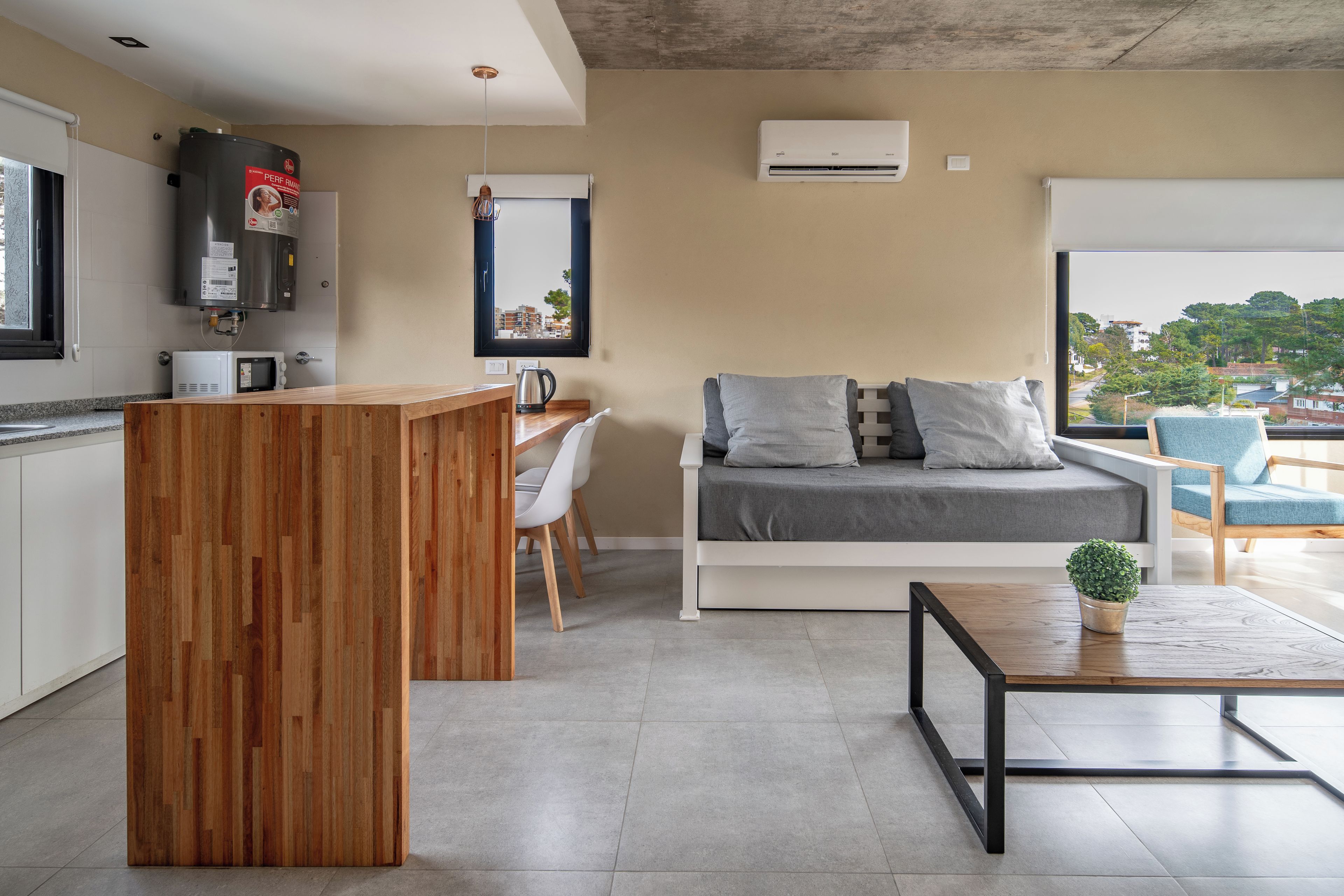 Una cocina moderna con una isla de madera, un comedor con mesa y sillas blancas, un sofá gris con cojines y vistas a la ciudad a través de un ventanal grande.