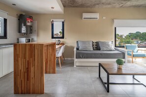 A modern kitchen with a wooden island, a dining area with a white table and chairs, a grey sofa with cushions, and a view of the city through a large window.