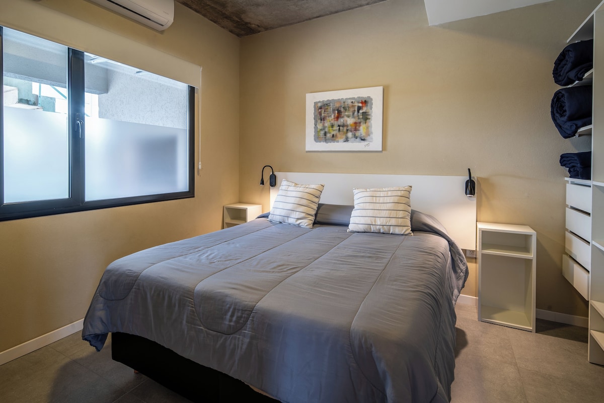 Ein Schlafzimmer mit einem Bett, Nachttischen, einer Wand-Klimaanlage, einem Fenster und einem Bild.