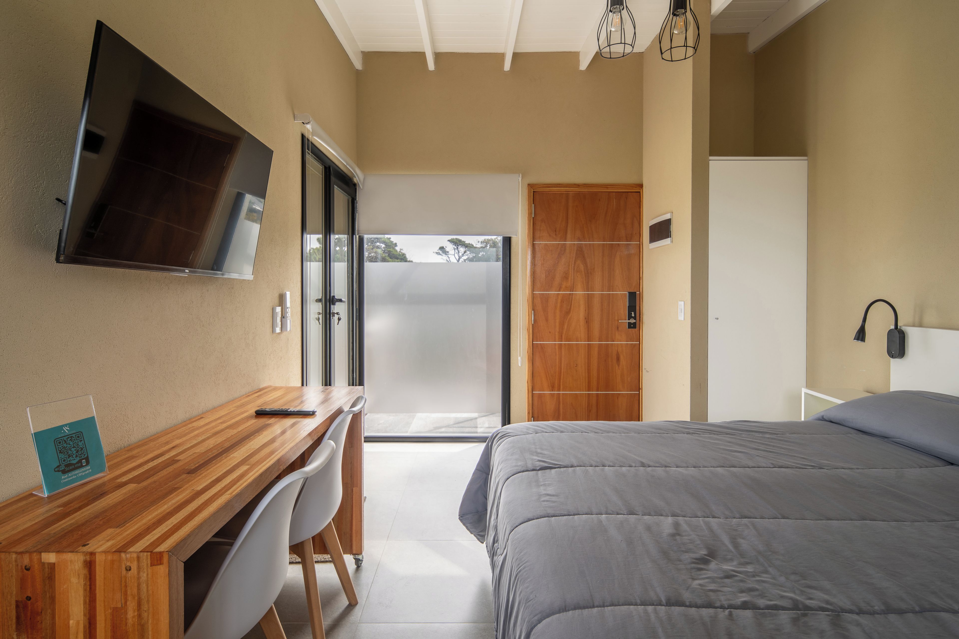 A bedroom with a bed, a desk with a chair, a TV mounted on the wall, and a door leading to a balcony.