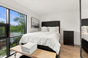 Ein modernes Schlafzimmer mit einem großen Bett, einem Nachttisch mit Handtüchern, einer Kommode und Blick auf Grün durch das Fenster.
