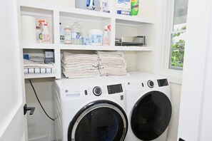 A laundry area with a front-loading washing machine and dryer, shelves with cleaning supplies, and a window with a view of greenery. - Private Estate with Pool & Two Pickleball Courts Directly on Twin Coves Beach (Governor's Harbour)