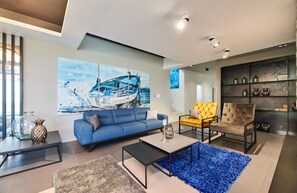 Ein modernes Wohnzimmer mit einem blauen Sofa, einem gelben Sessel und einem Couchtisch. An der Wand hängt ein großes Gemälde eines Bootes und eine Bücherwand ist mit dekorativen Objekten geschmückt.