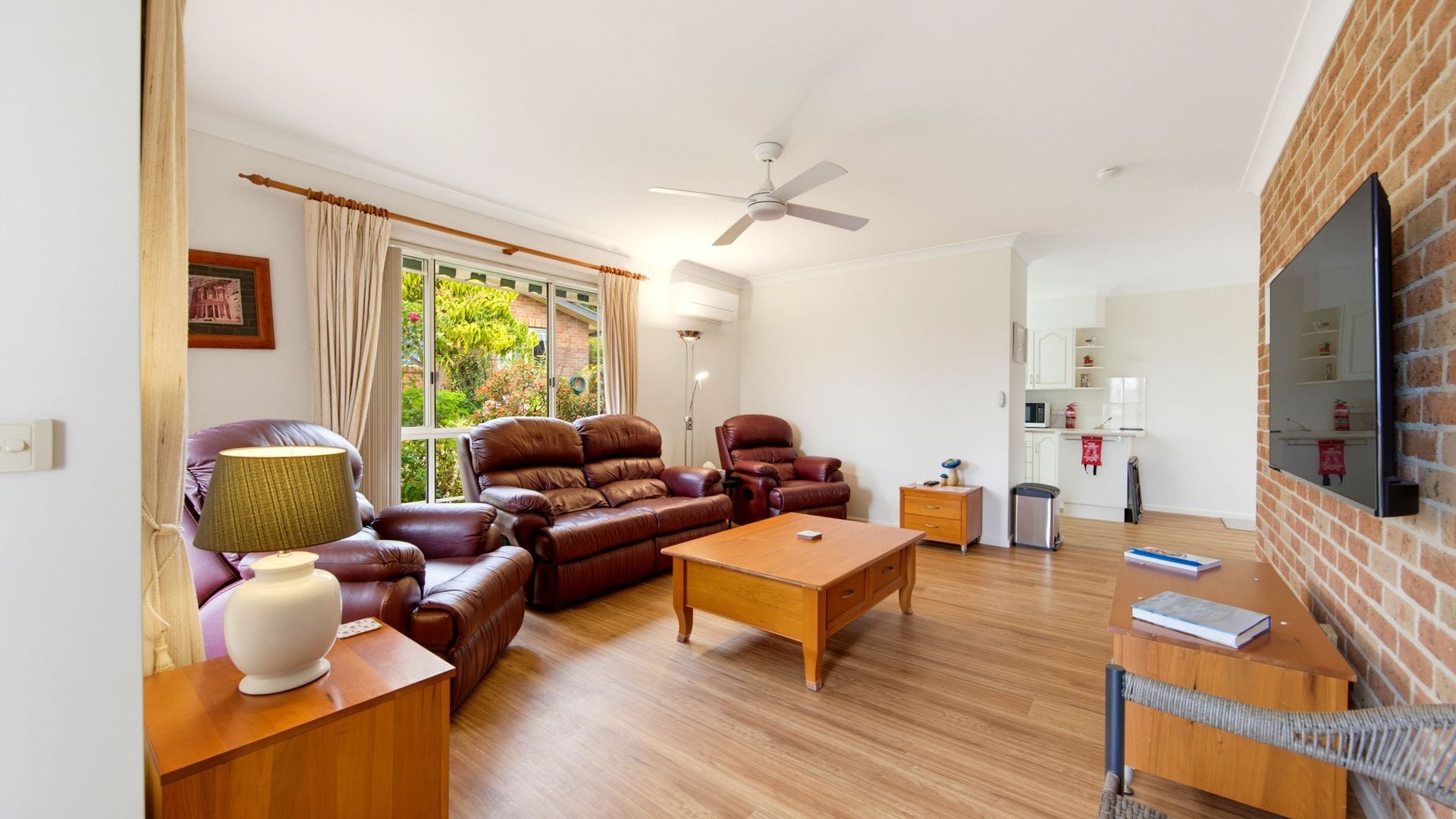 A living room with leather sofas, a wooden coffee table, a floor lamp, and a television mounted on a brick wall.