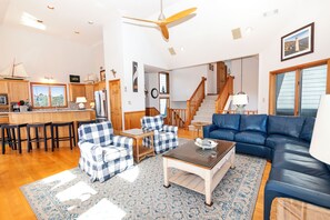 Ein geräumiges Wohnzimmer mit einem blauen Sofa, karierten Sesseln, einem Couchtisch und einer Küchenzeile mit Holzschränken und Blick durch das Fenster.