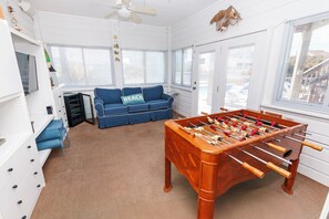 Et værelse med et fodboldbord, en blå sofa med pude med teksten 'BEACH' og en loftsvifte.