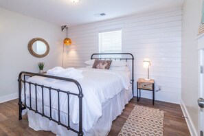 A bedroom with a metal bed frame, white bedding, a small bedside table with a lamp, a round mirror on the wall, and a window with blinds. - Free 6-Seat Golf Cart | Private Free Heated Pool | Walk to Beach | Sleeps 18 (Hilton Head Island)