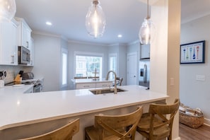 A modern kitchen with white cabinetry, a central island with seating, pendant lighting, and a view of the outside. - Free 6-Seat Golf Cart | Private Free Heated Pool | Walk to Beach | Sleeps 18 (Hilton Head Island)