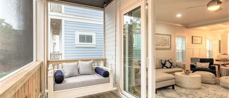 A porch with a swing seat, a sliding glass door leading to a living room with a sofa and ottoman, and a wooden deck.