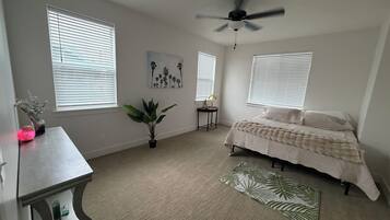 A bedroom with a bed, a ceiling fan, two windows with blinds, a small table with a plant, and a decorative rug.