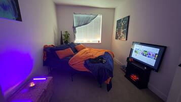 A bedroom with a bed, a television, a window with blinds, and a wall-mounted shelf.
