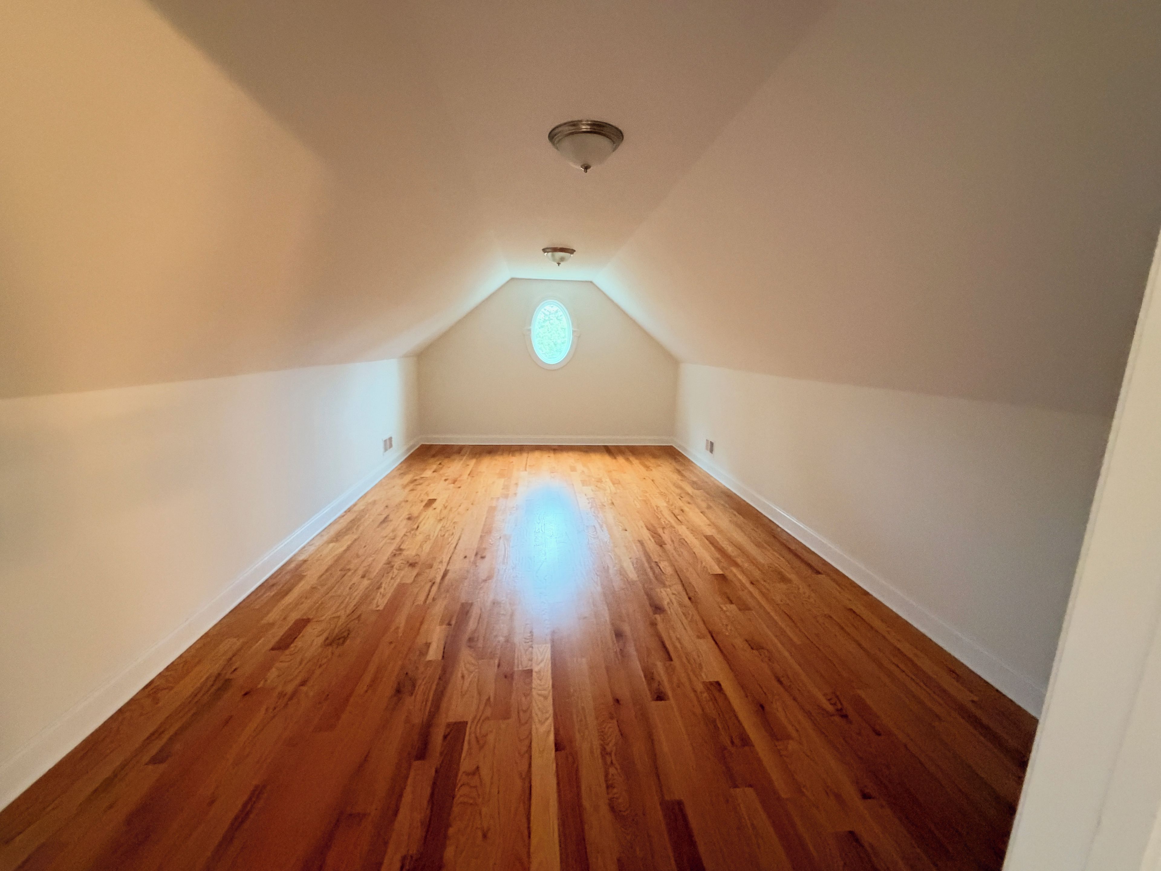 An attic room with wooden flooring, white walls, and a sloped ceiling.