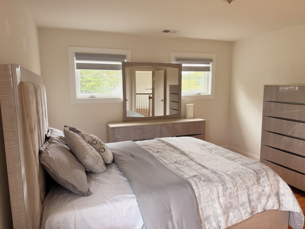 Un dormitorio con cama, cómoda, espejo y dos ventanas.