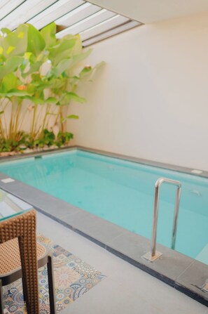 An indoor swimming pool with a clear view of the water, a metal pool ladder, and a wicker chair.