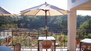 A balcony with a table and chairs, a large umbrella, and a view of a forested landscape.