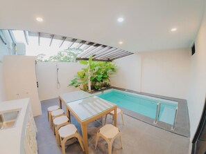 A modern outdoor pool area with a glass table and chairs, surrounded by plants and a pergola.