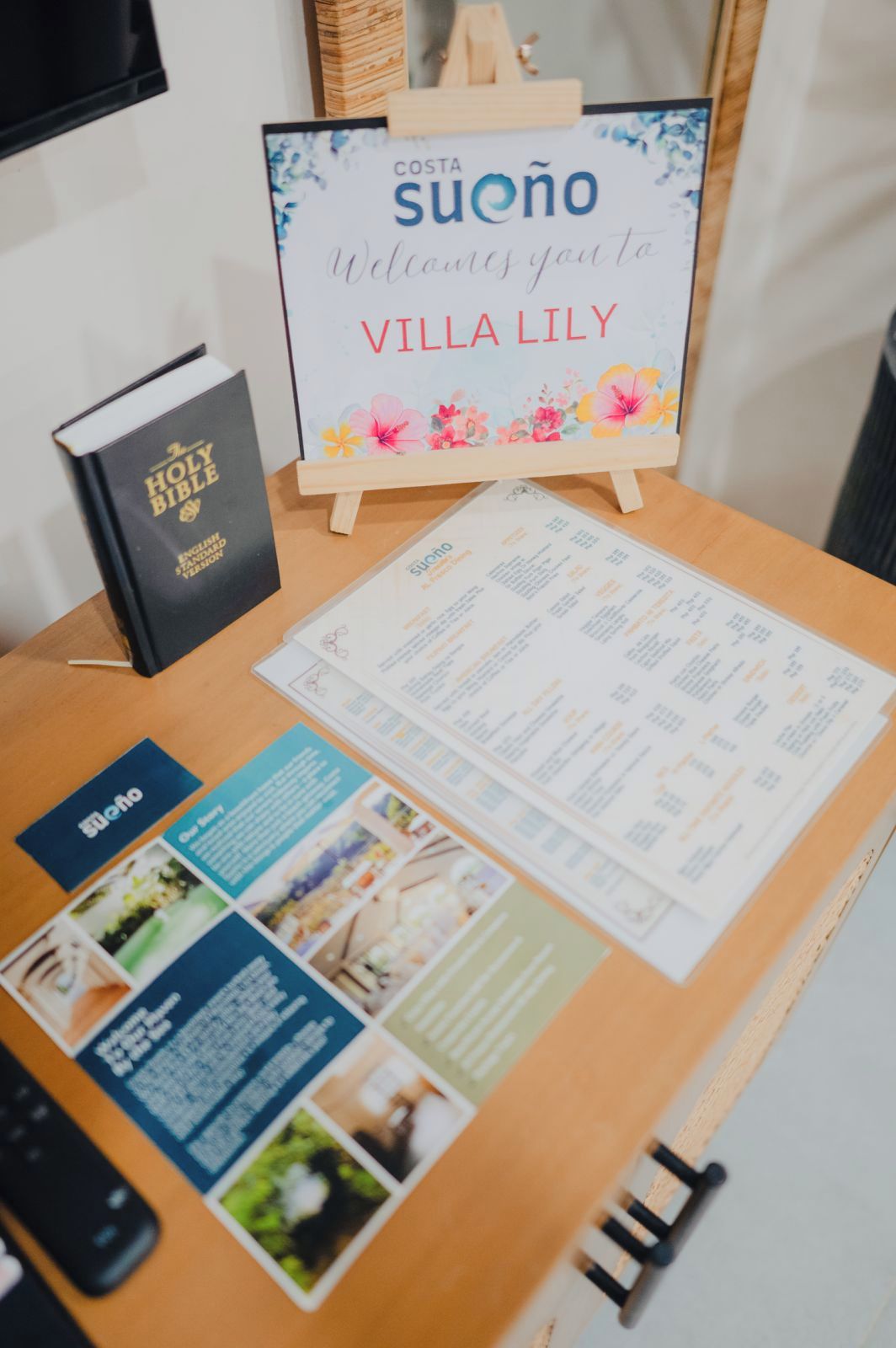 A wooden table with a welcome sign, a Holy Bible, a menu, and a remote control.