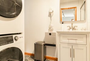 A bathroom with a washing machine, a hairdryer, a trash bin, and a sink with a mirror. - Fully Renovated, King Bed, Dog Paradise, 4-Acres (Sidney Center)