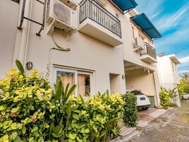 A residential building with a balcony, a car parked in the driveway, and lush greenery in the foreground.