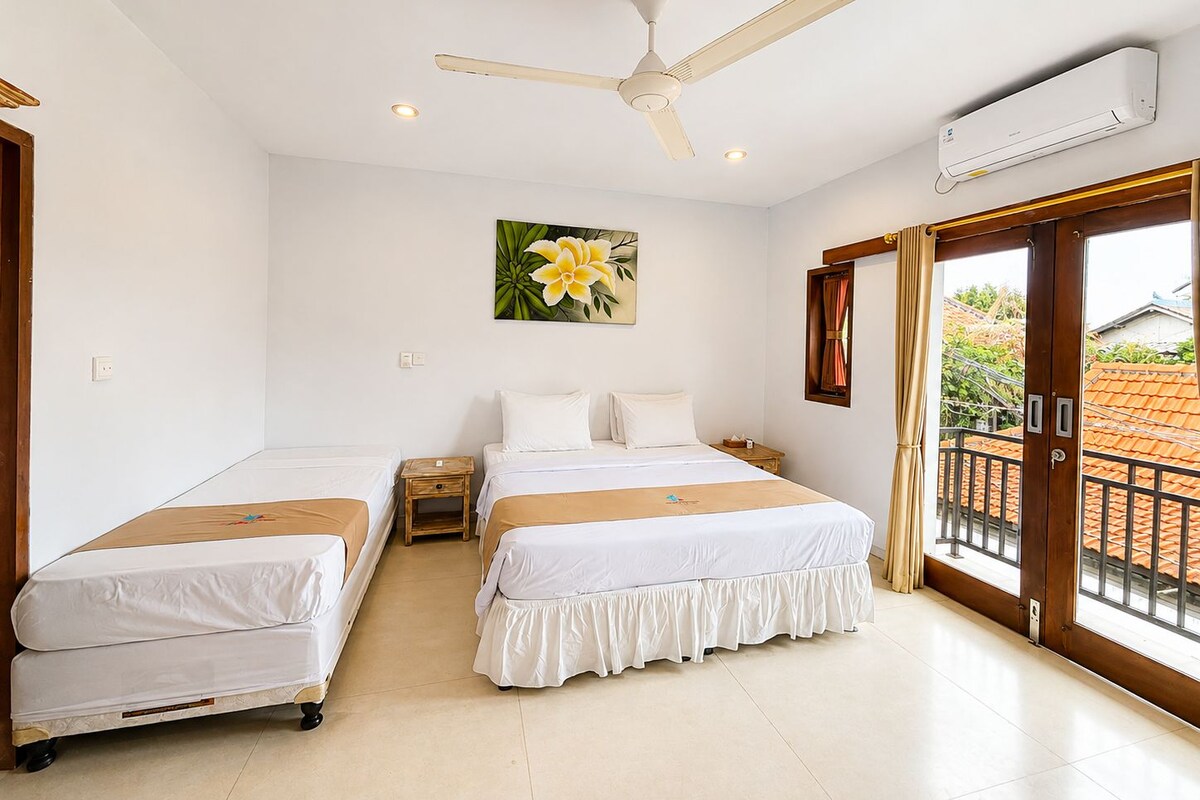 Une chambre d’hôtel avec deux lits, un ventilateur de plafond, un appareil de climatisation, un tableau représentant une fleur et une vue sur l’extérieur.