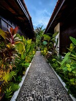 A pathway flanked by lush tropical plants and wooden buildings with a tiled floor.