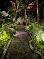 A stone pathway flanked by lush tropical plants leading to a wooden structure.
