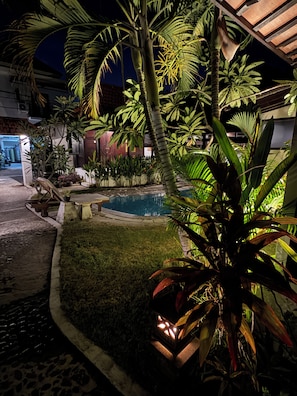 A nighttime view of a courtyard with a swimming pool, surrounded by various plants and a building with a covered entrance. - Surfer Paradise Villas Bali (Pererenan)