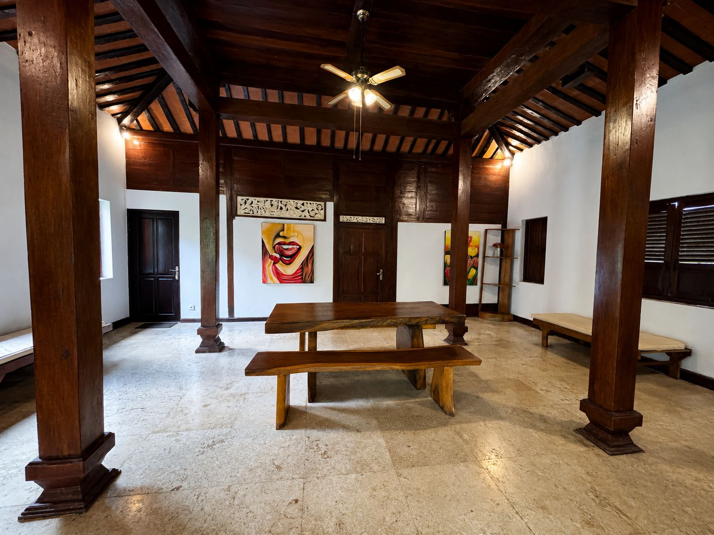 Una habitación amplia con techo de madera, ventilador de techo, mesa de madera y obras de arte en las paredes.
