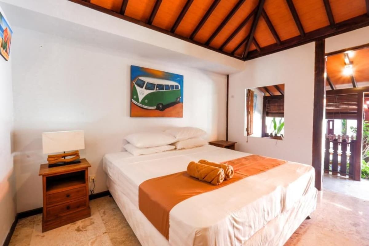 A bedroom with a bed, a nightstand, a painting of a green van, and a window with blinds.