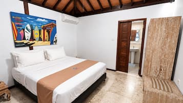 A bedroom with a bed, a painting, an air conditioning unit, a wooden door, and a bathroom visible through an open door.