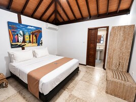 A bedroom with a bed, a painting, an air conditioning unit, a wooden door, and a bathroom visible through an open door.