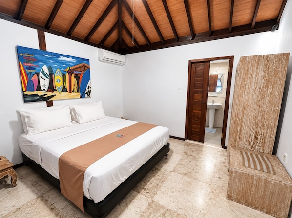 A bedroom with a bed, a painting, an air conditioning unit, a wooden door, and a bathroom visible through an open door. - Surfer Paradise Villas Bali (Pererenan)