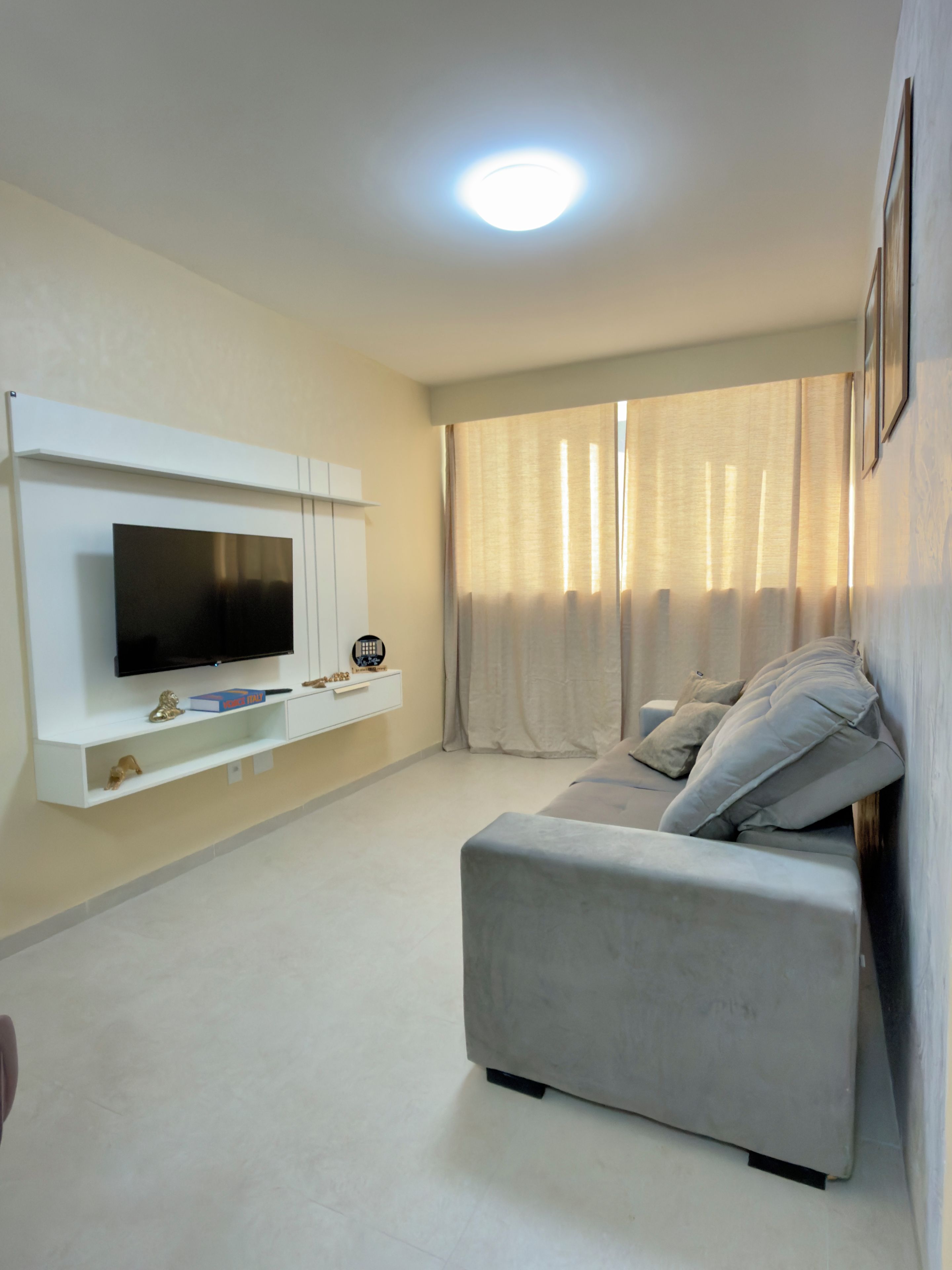 Een moderne woonkamer met een flatscreen televisie aan een witte muur, een grijze bank en een groot raam met gordijnen.