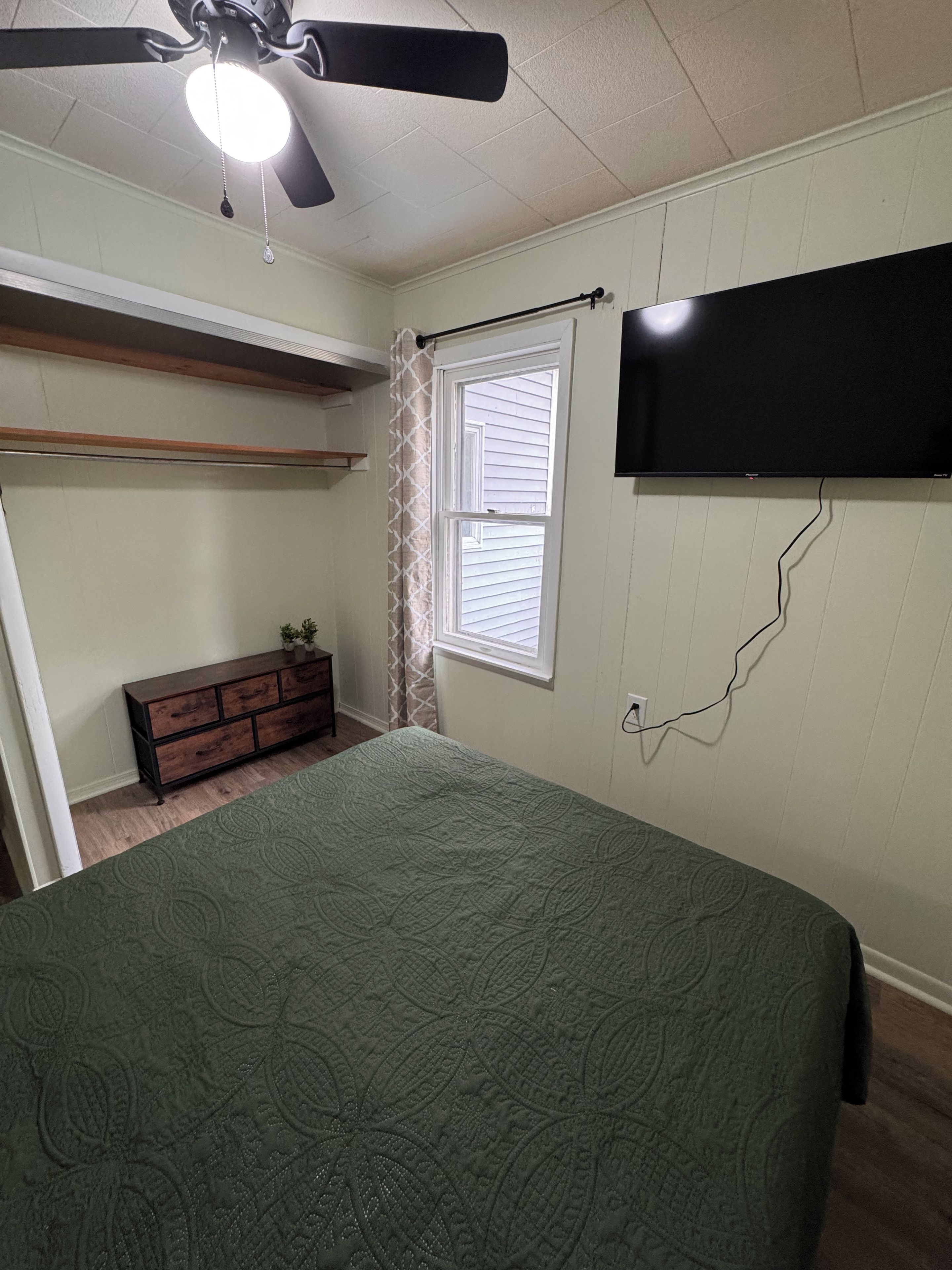 Een slaapkamer met een bed, een plafondventilator, een raam, een dressoir en een aan de muur bevestigde televisie.