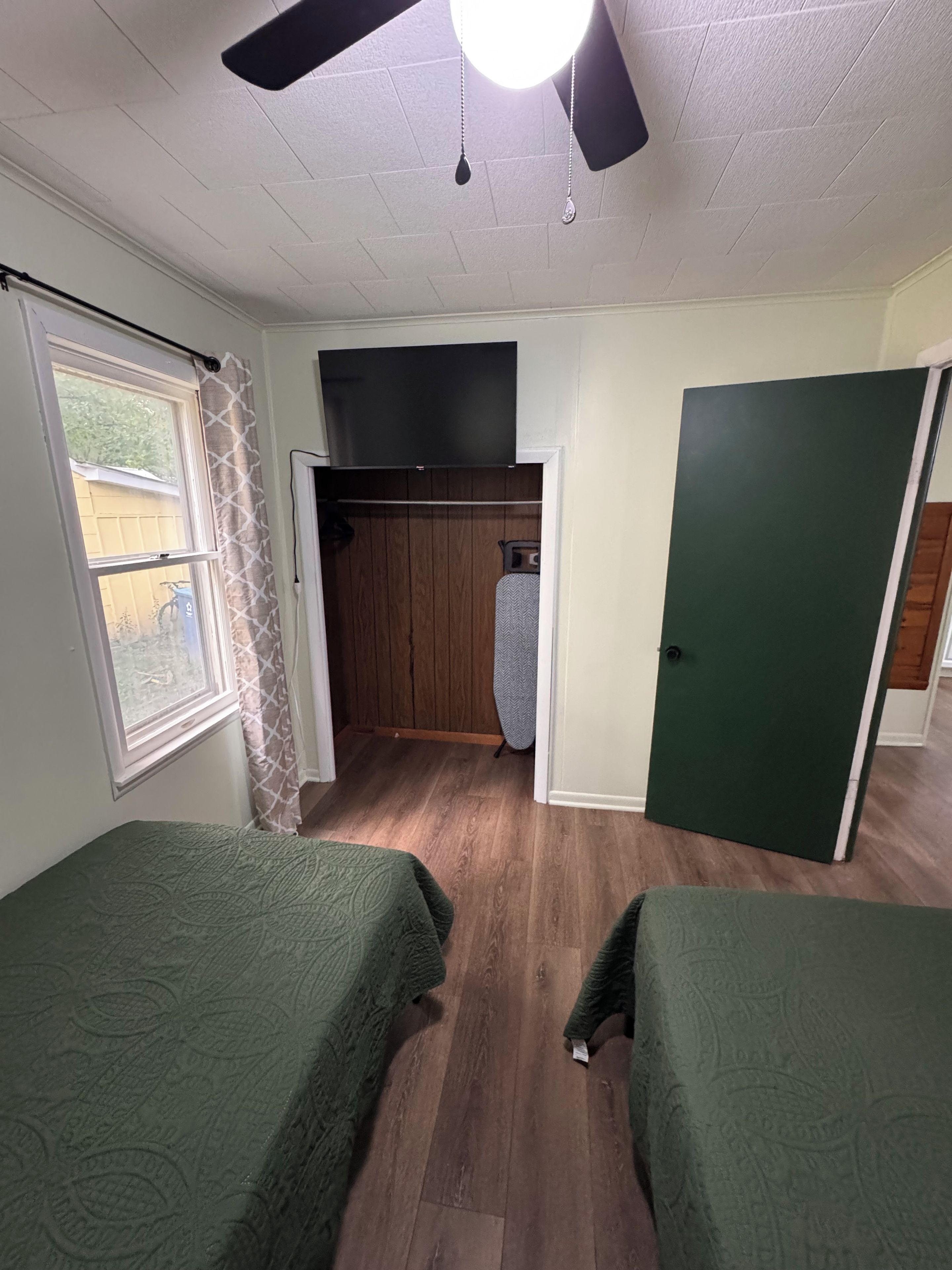 A room with two beds, a ceiling fan, a window with curtains, a door, and a small closet with a mirror.