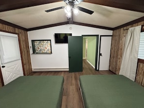 A room with two beds, a ceiling fan, a wall-mounted TV, and a door leading to another room.