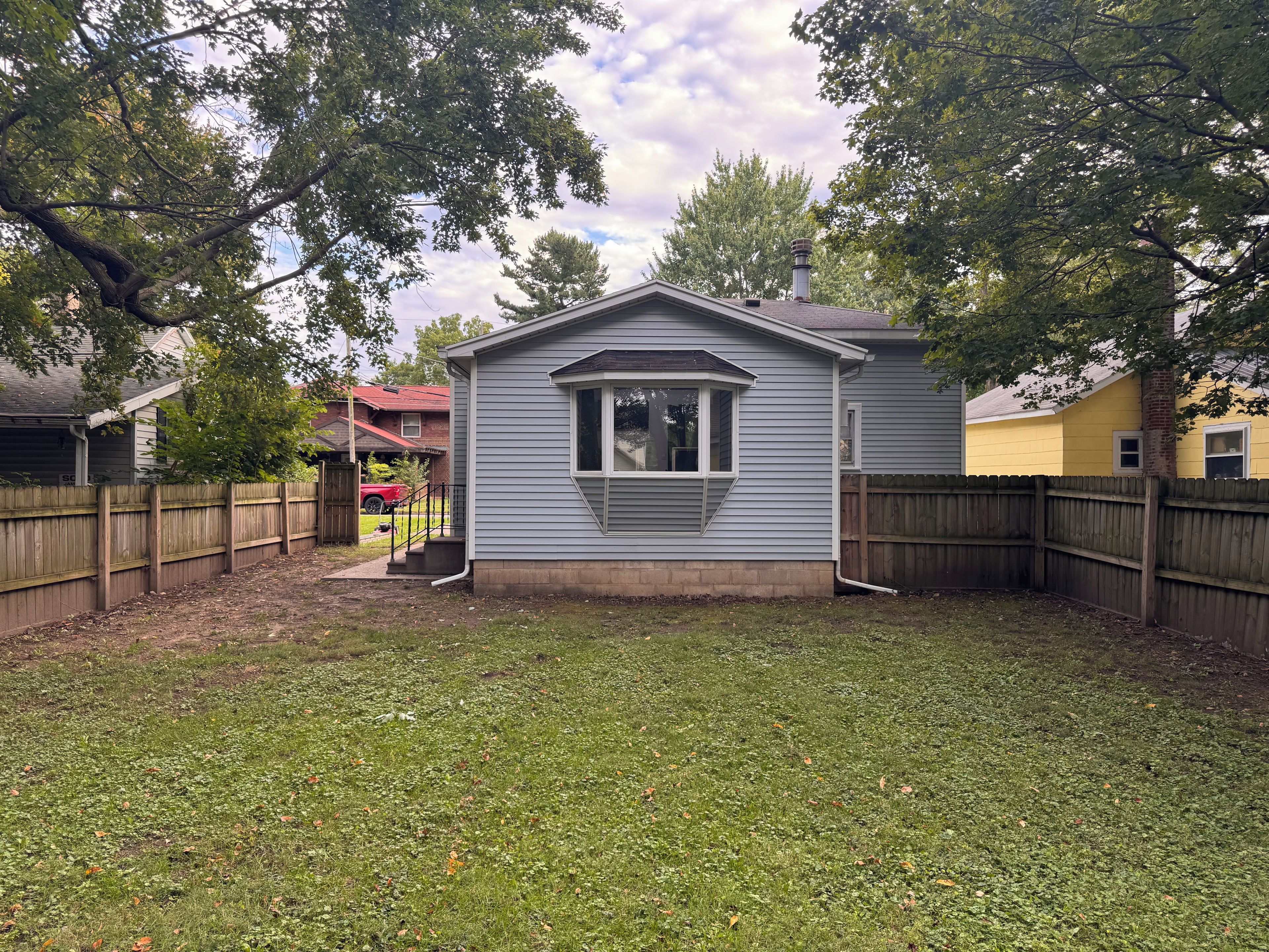 A small, single-story house with a fenced backyard, a chimney, and a red vehicle parked in the background.