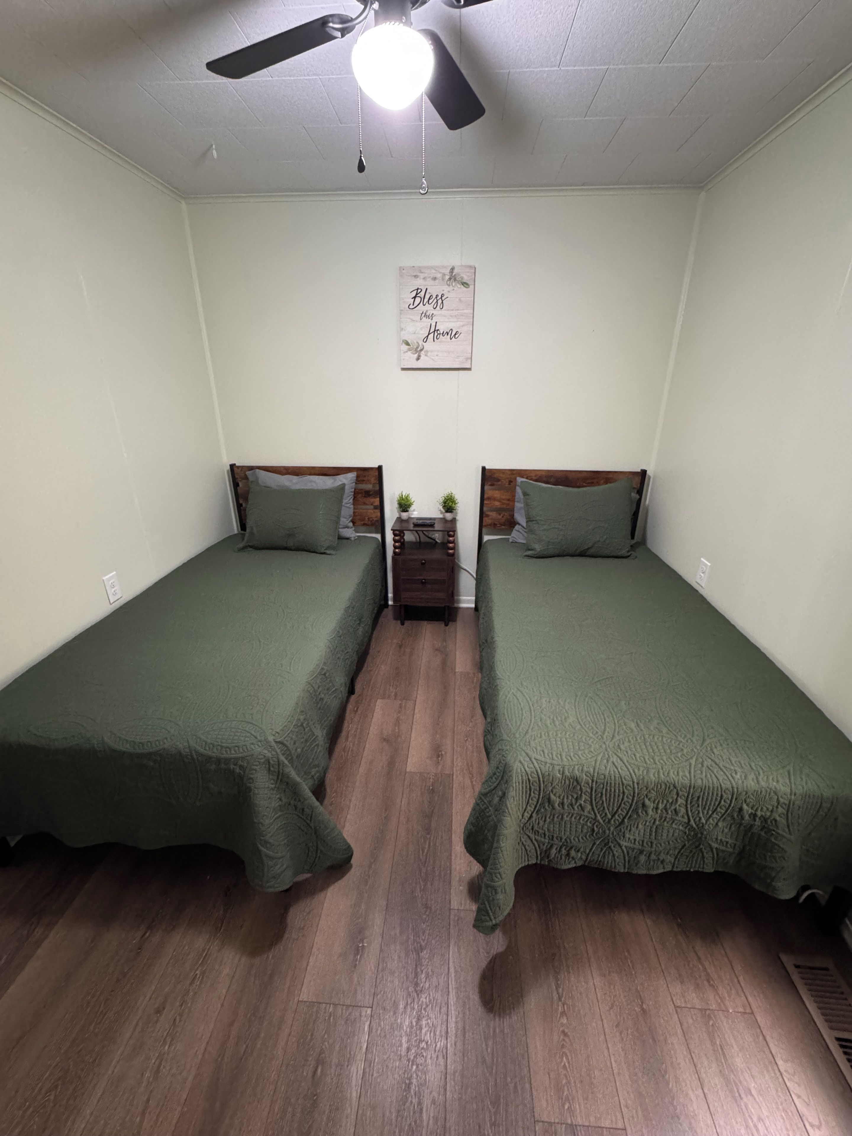 A room with two single beds, a ceiling fan, a small nightstand with a plant, and a wall sign.