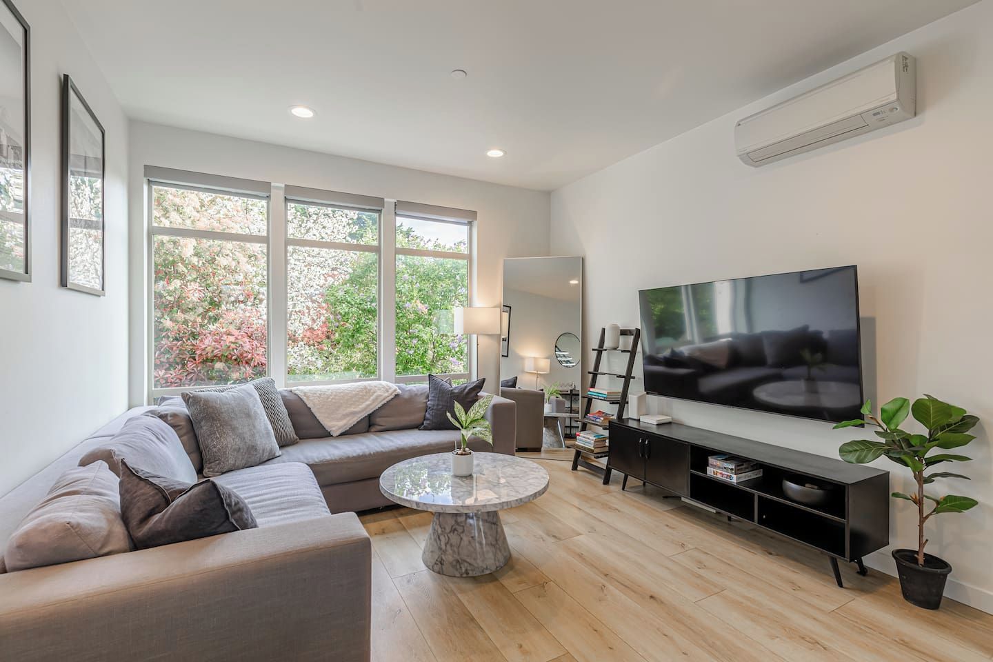 A modern living room with a sectional sofa, a flat-screen TV, a coffee table, and a large window with a view of trees.