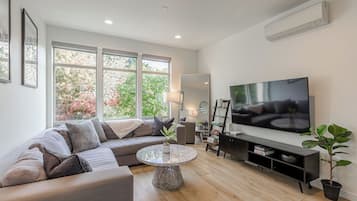 A modern living room with a sectional sofa, a flat-screen TV, a coffee table, and a large window with a view of trees.