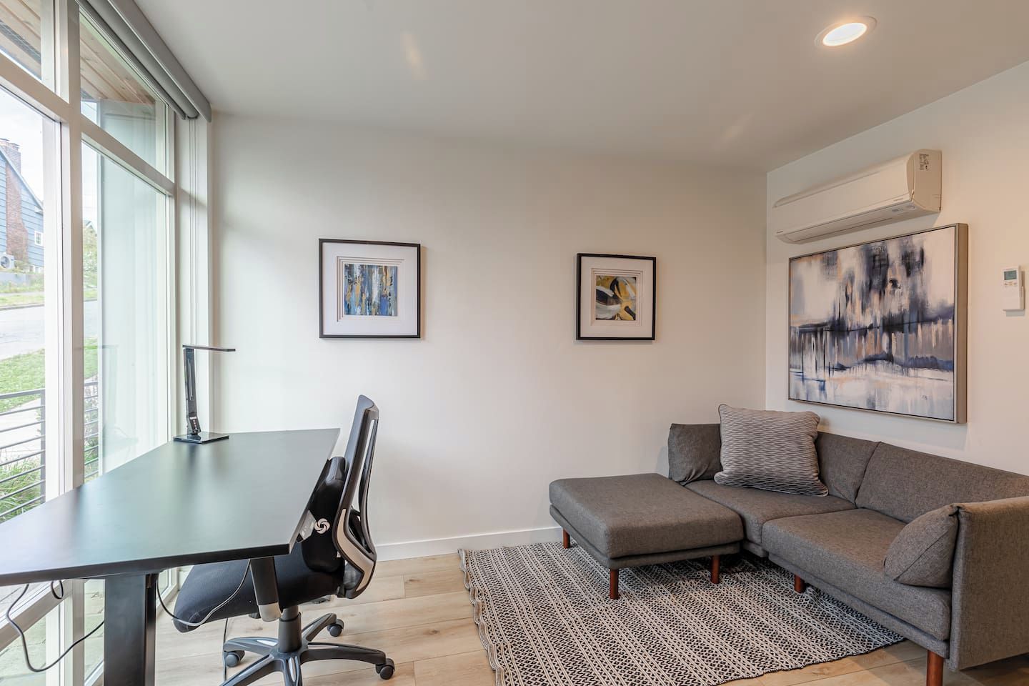 Un salon moderne avec un bureau, un canapé gris et deux œuvres d’art encadrées accrochées au mur.