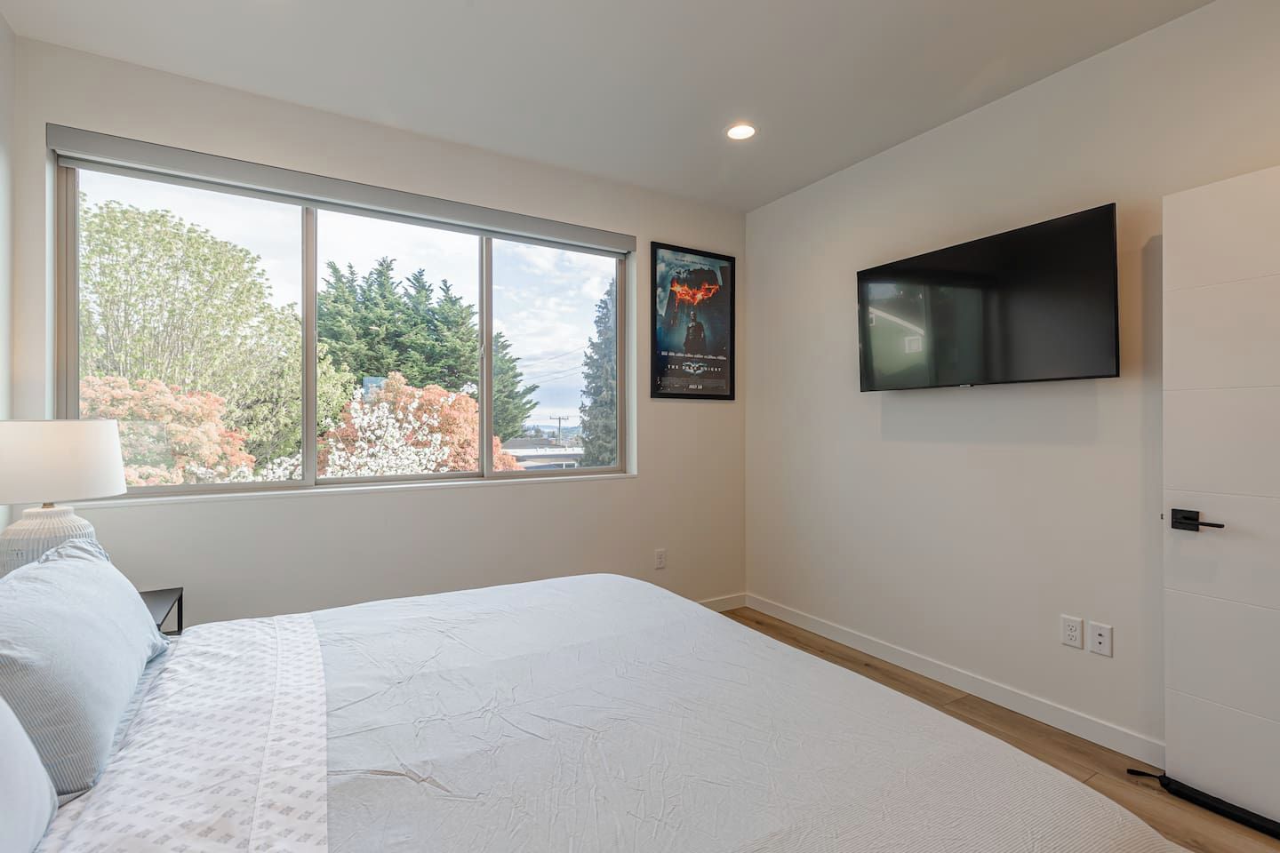 Ein Schlafzimmer mit großem Fenster, einem Bett, einem an der Wand befestigten Fernseher und einem gerahmten Bild.
