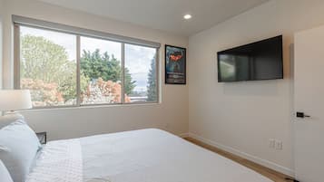 Ein Schlafzimmer mit großem Fenster, einem Bett, einem an der Wand befestigten Fernseher und einem gerahmten Bild.