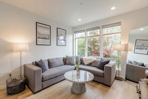 Un salon moderne avec un canapé d’angle gris, une table basse en marbre et de grandes fenêtres donnant sur la verdure.
