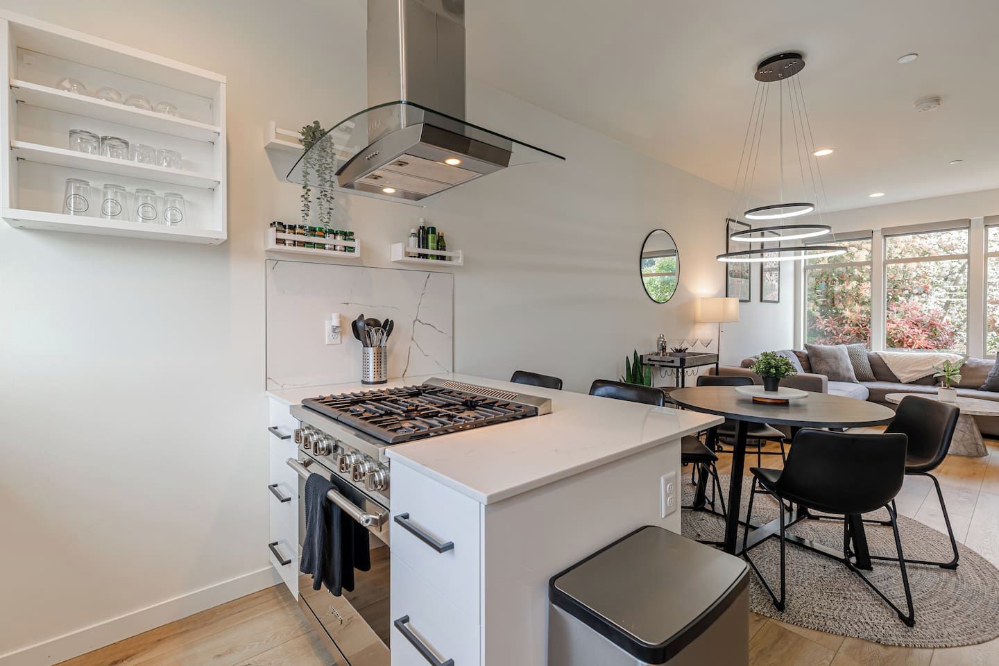 Cozinha moderna com ilha central, área de refeições com mesa redonda e sala de estar com sofá e vista para o exterior.