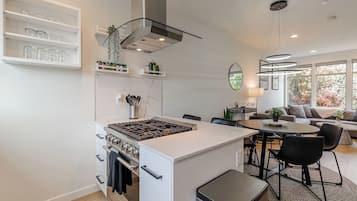 Cozinha moderna com ilha central, área de refeições com mesa redonda e sala de estar com sofá e vista para o exterior.