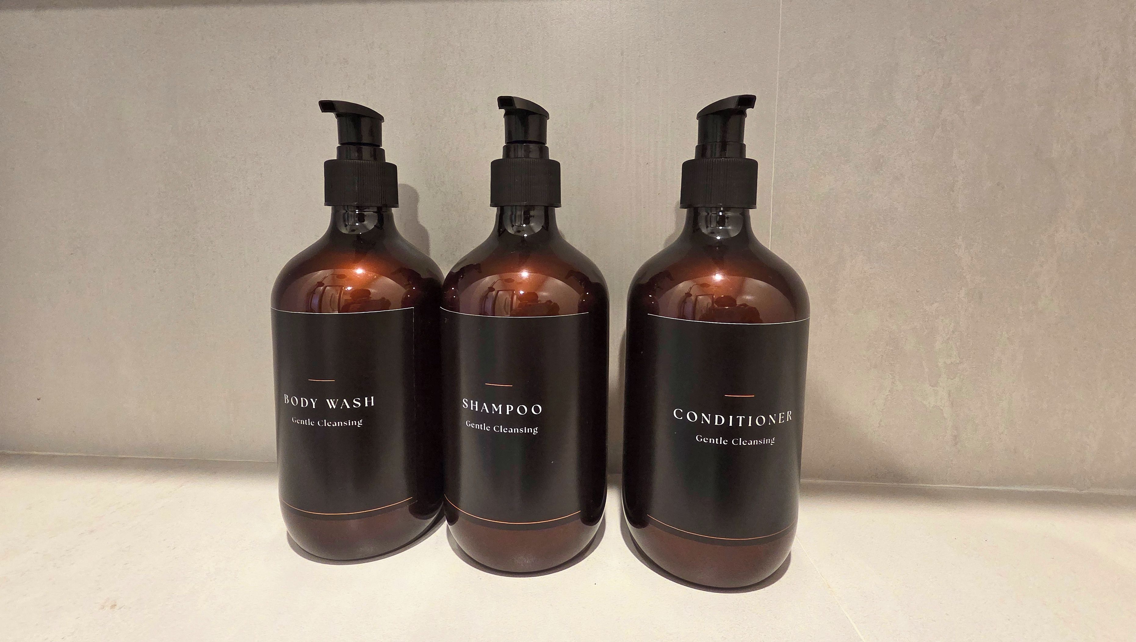 Trois bouteilles de gel douche, de shampoing et d’après-shampoing sur une surface blanche.
