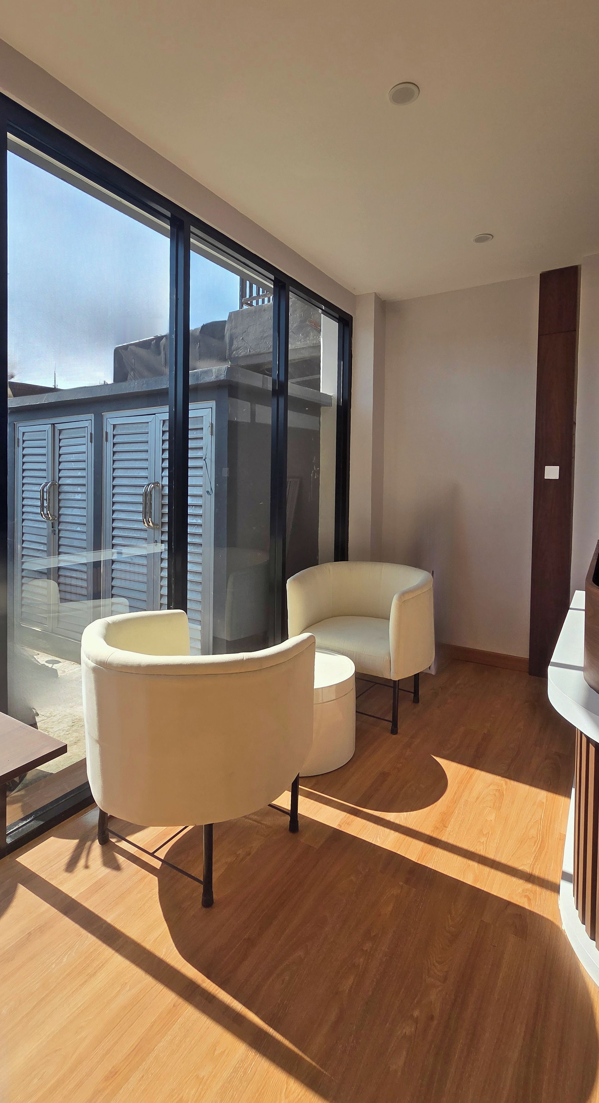 Una sala de estar moderna con un ventanal grande, dos sillones blancos, un pequeño reposapiés y piso de madera.