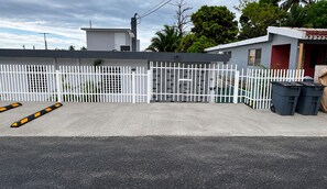A gated parking area with a white fence, a building with a red door, and two black trash bins. - EncantoPlayeroPR is the choice you should make to stay comfortable (Playa Fortuna)